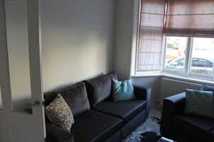 A living room with a dark grey sofa, patterned cushions, a window with a view of parked cars, and a radiator.