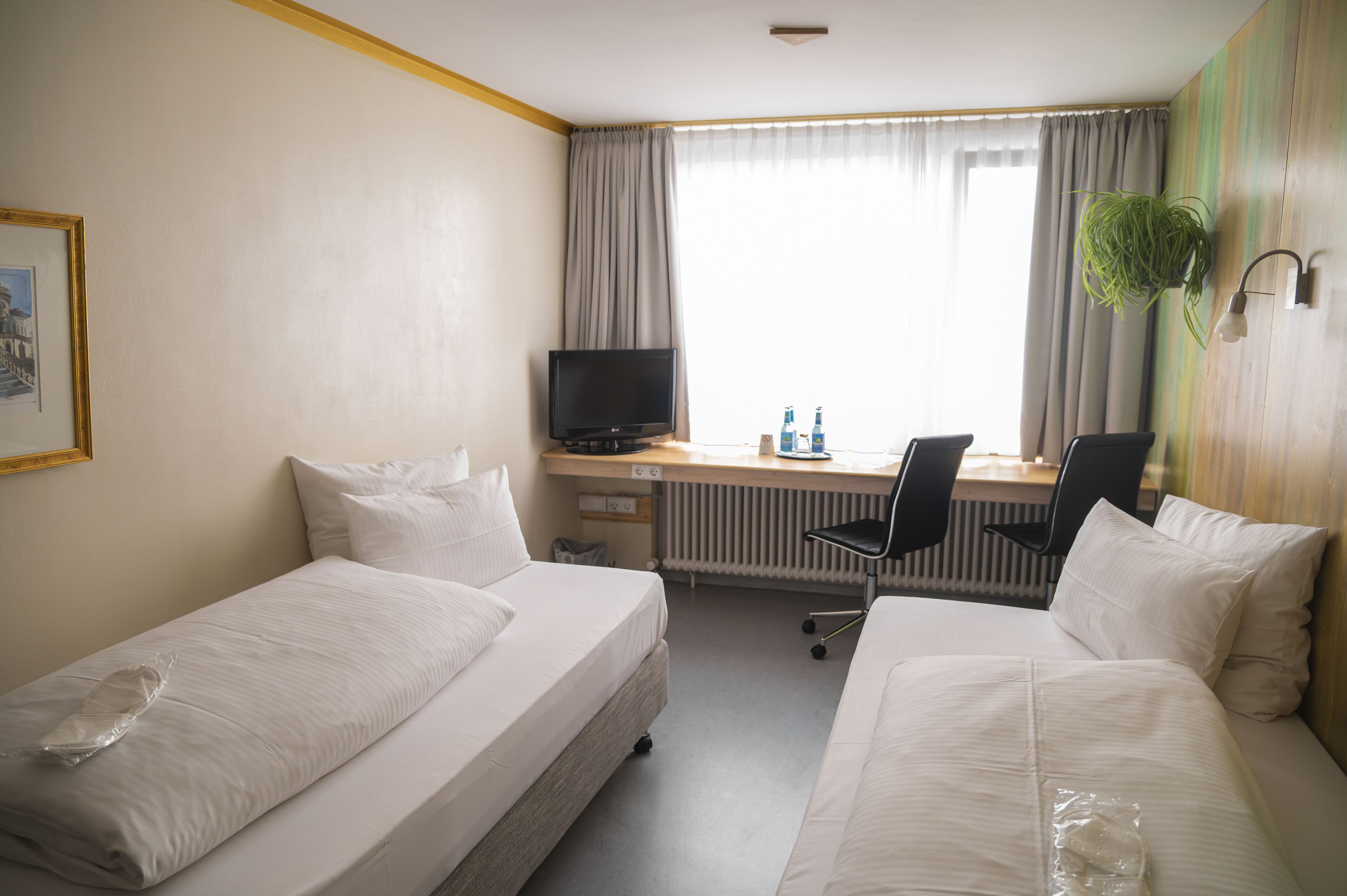 Habitación de hotel con dos camas individuales, un escritorio con computadora y dos sillas, televisor y ventana con cortinas.