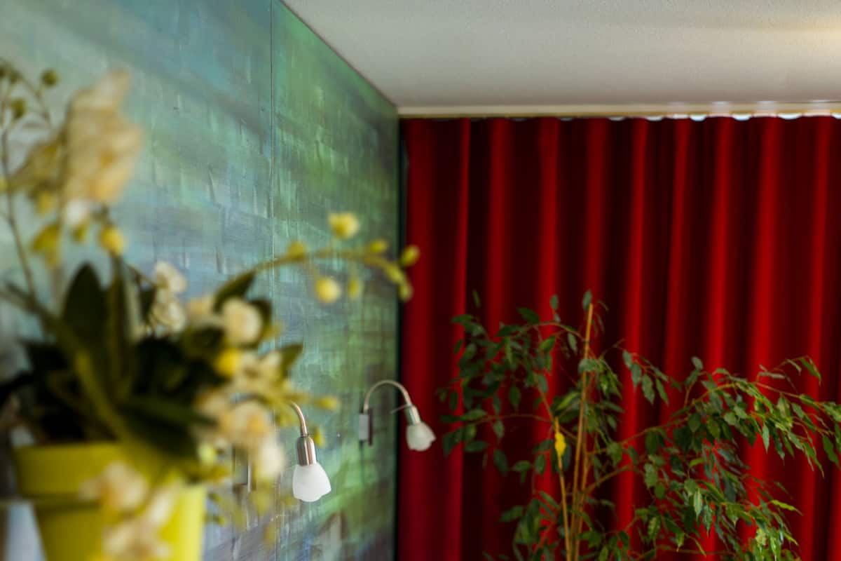 Una cama bien hecha con sábanas y almohadas blancas, una mesita de noche con una lámpara, una planta en maceta y una cortina roja al fondo.