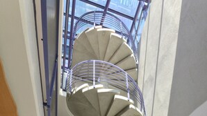Una escalera espiral de varios niveles con barandillas de metal y base de hormigón, rodeada de guijarros blancos y flores rojas.