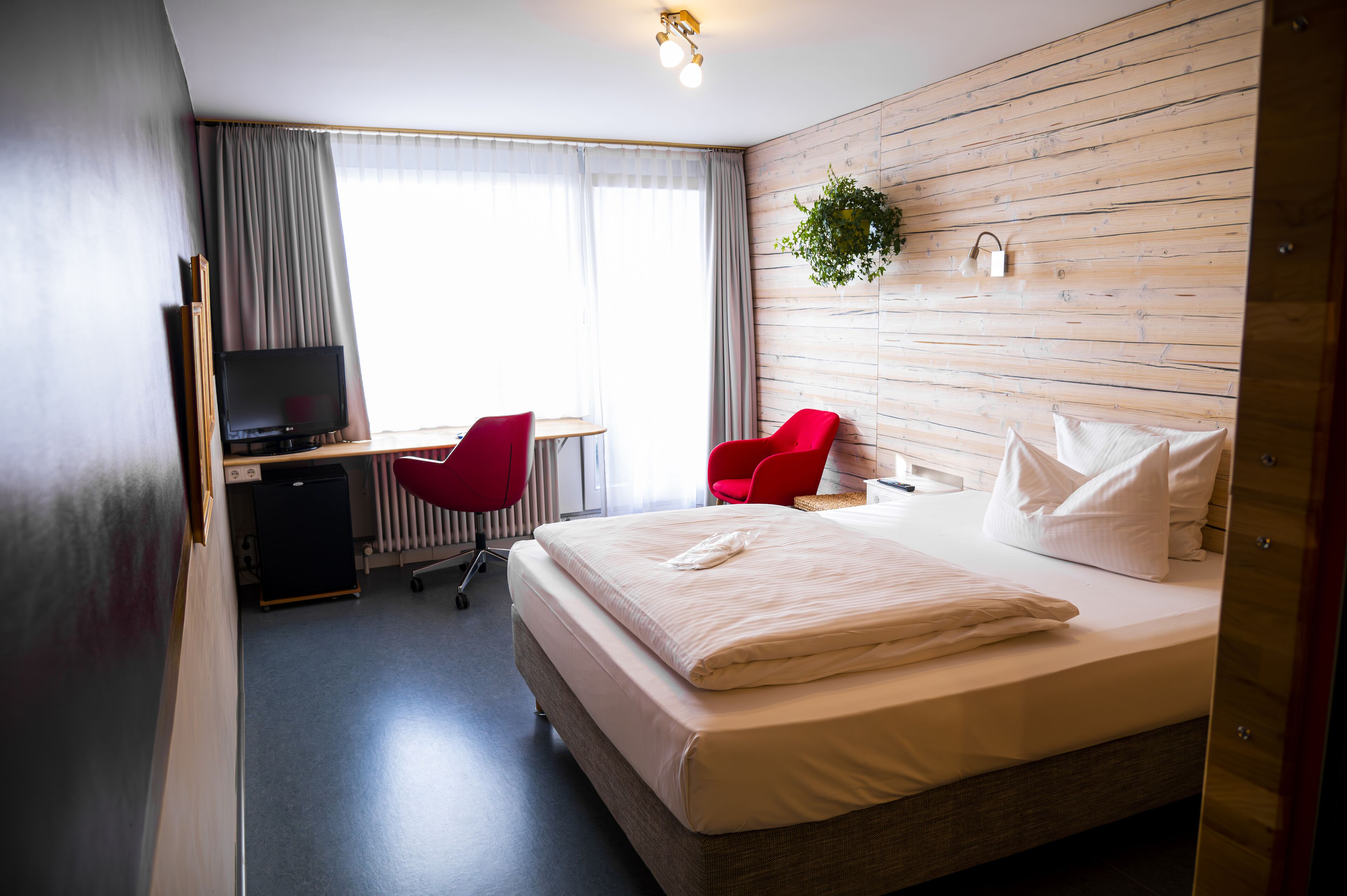 Ein Hotelzimmer mit einem Bett, einem Schreibtisch mit Computer, zwei roten Sessel, einem Fernseher und einer Holzvertäfelung.