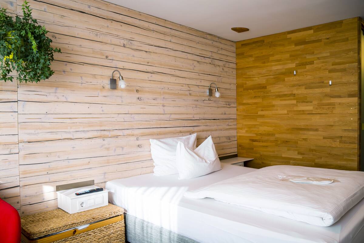 Una habitación de hotel moderna con una pared de madera, una cama con ropa blanca, una mesita de noche con una lámpara y una silla roja.