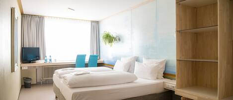 A hotel room with a bed, a desk with a computer, a chair, a television, and a window with curtains.
