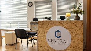 Una recepción con un letrero que dice "CENTRAL GUEST HOUSE" en un interior moderno con suelo de madera, un mostrador, sillas y una planta.
