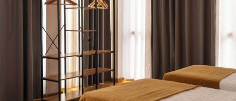A hotel room with a wooden floor, two beds with brown bedding, a metal shelving unit with clothes hanging, and a window with curtains.