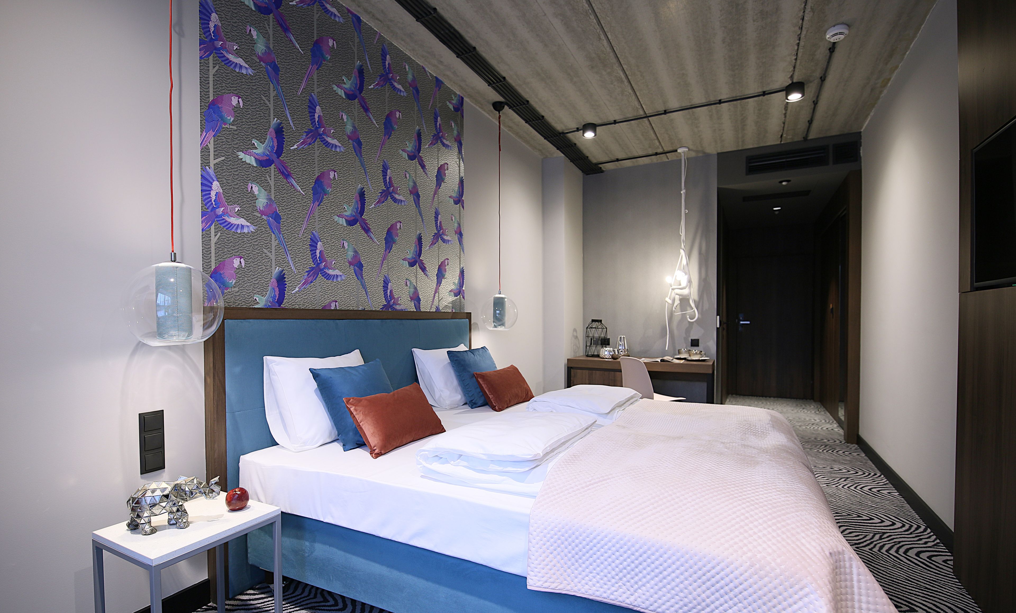 Une chambre d’hôtel moderne avec un grand lit, une table de chevet, un miroir et un mur orné d’un motif représentant des oiseaux.