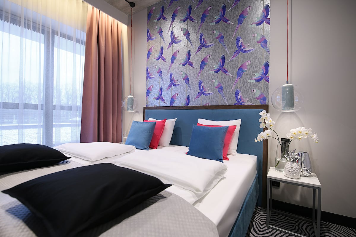 Quarto de hotel com uma cama grande, uma mesa de cabeceira, parede com padrão de pássaros e janela com cortinas.