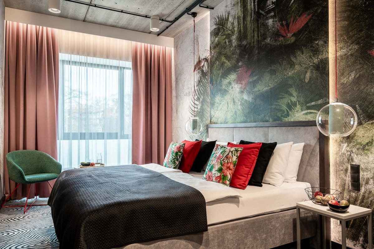 Quarto de hotel moderno com uma cama grande, uma cadeira verde, uma mesinha com frutas e um quadro abstrato de grandes dimensões na parede.