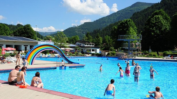 A swimming pool with a rainbow slide, surrounded by mountains and trees. - Hotel Restaurant Pension Krone Post (Simonswald)