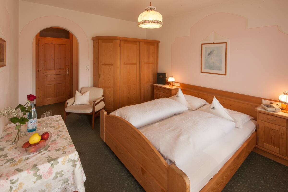 A bedroom with two wooden beds, a wooden wardrobe, a bedside table with a lamp, and a floral tablecloth.