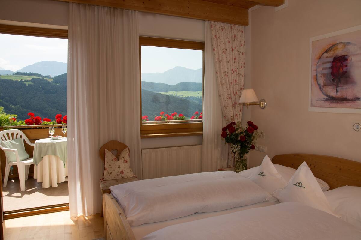 A bedroom with a bed, a window with a view of mountains, a table with a chair, and a vase with flowers.