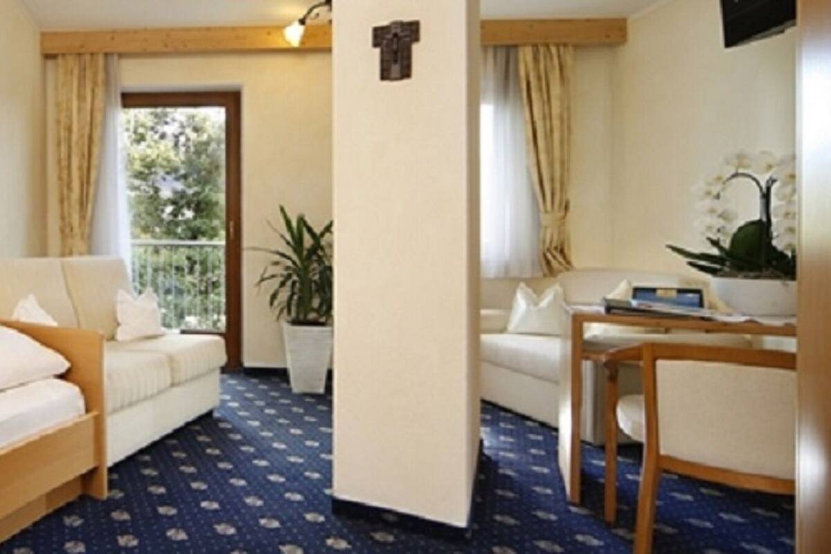 A hotel room with a bed, a sofa, a desk with a chair, a potted plant, and a window with curtains.