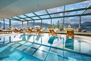 A large indoor swimming pool with clear blue tiles, surrounded by wooden lounge chairs and potted plants, with a view of mountains through large windows. - Berglandhotel Untertheimerhof (Villandro)