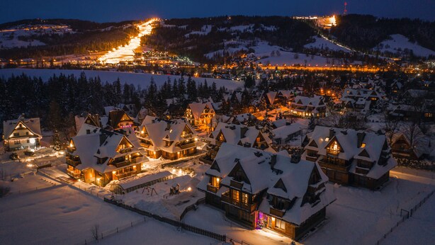 A snow-covered village with traditional wooden houses, illuminated by warm lights, and a distant ski resort in the background. - Apartamenty Sun&Snow Lipki Park Zakopane (Zakopane)