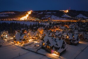 A snow-covered village with traditional wooden houses, illuminated by warm lights, and a distant ski resort in the background. - Apartamenty Sun&Snow Lipki Park Zakopane (Zakopane)