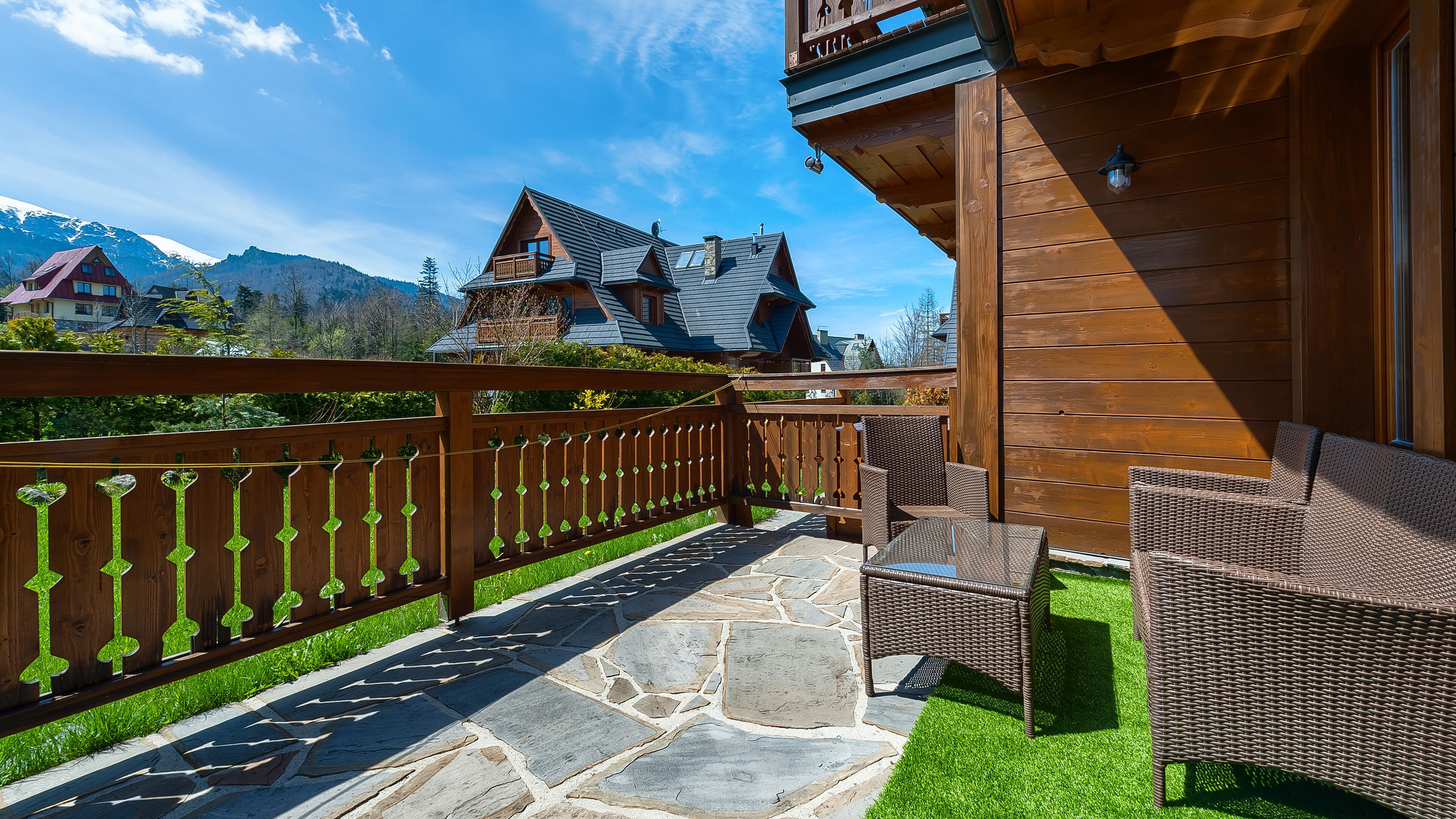 A wooden balcony with wicker furniture, stone flooring, and a view of a mountain house.