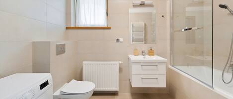 A compact bathroom with a toilet, washing machine, radiator, sink, and glass shower enclosure.