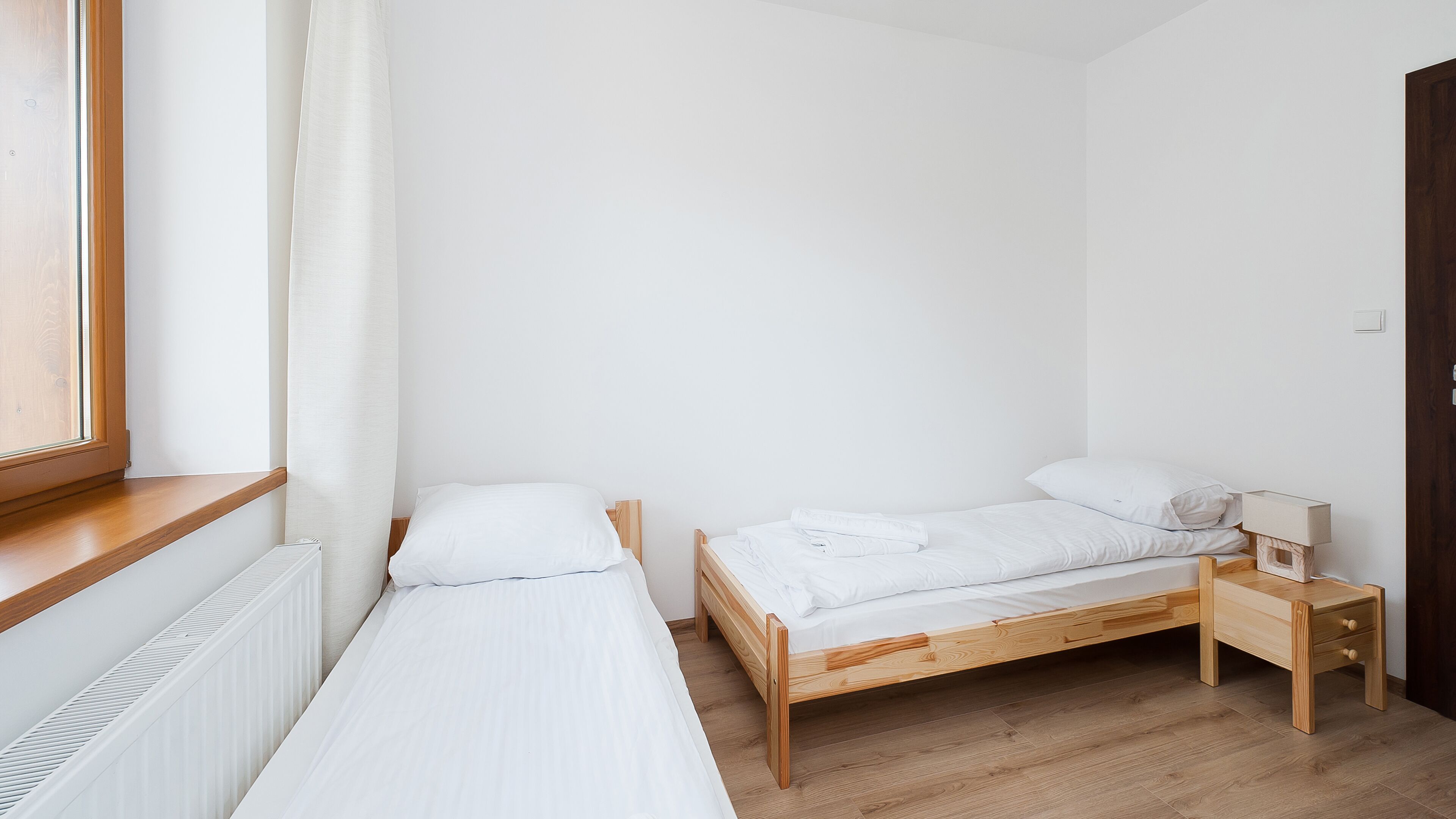 A small, clean room with two single beds, a wooden nightstand, a radiator, and a window with a curtain.
