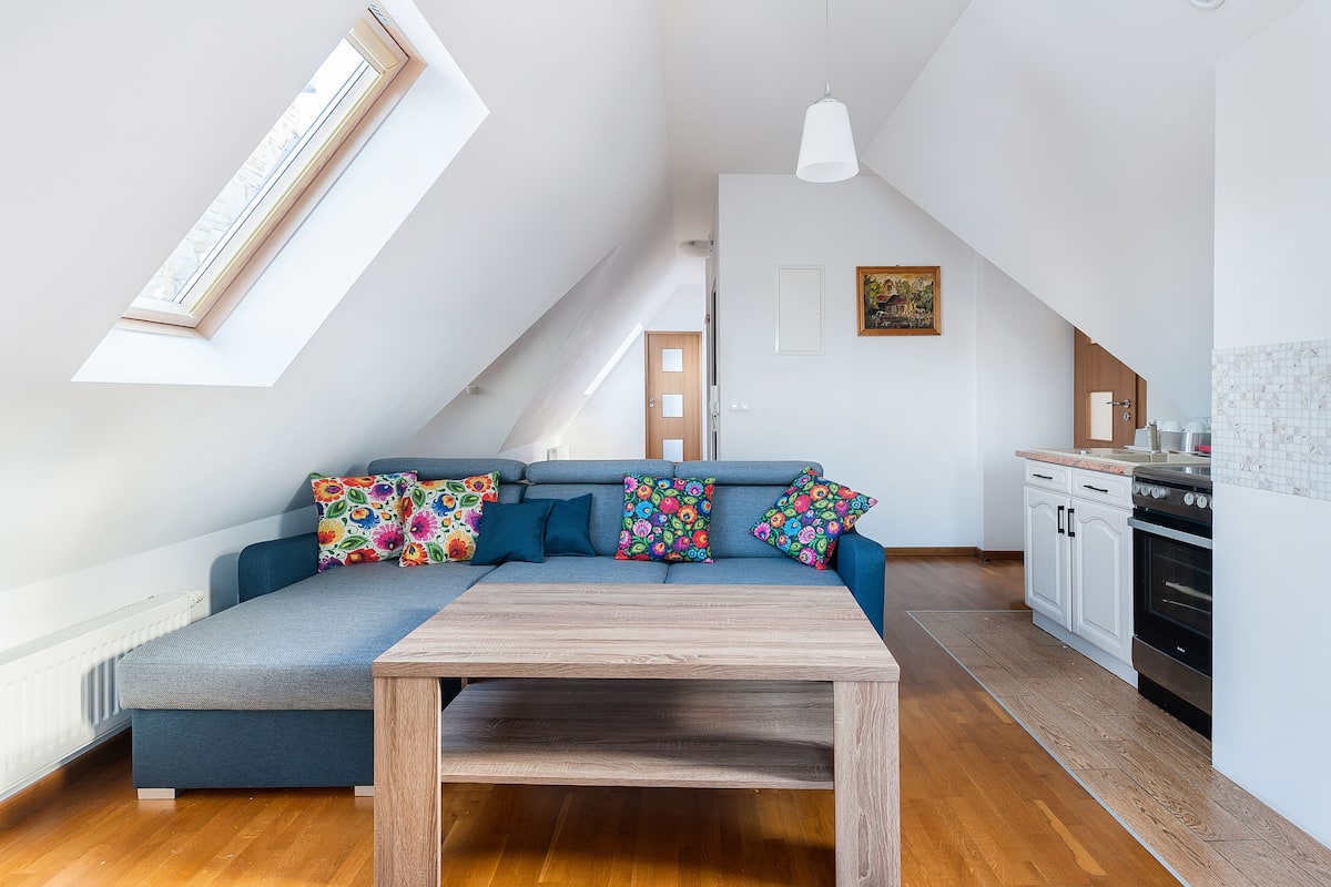 Un loft moderno con cama elevada, una mesa de comedor, una cocina con armarios blancos y un gran ventanal que proporciona luz natural.