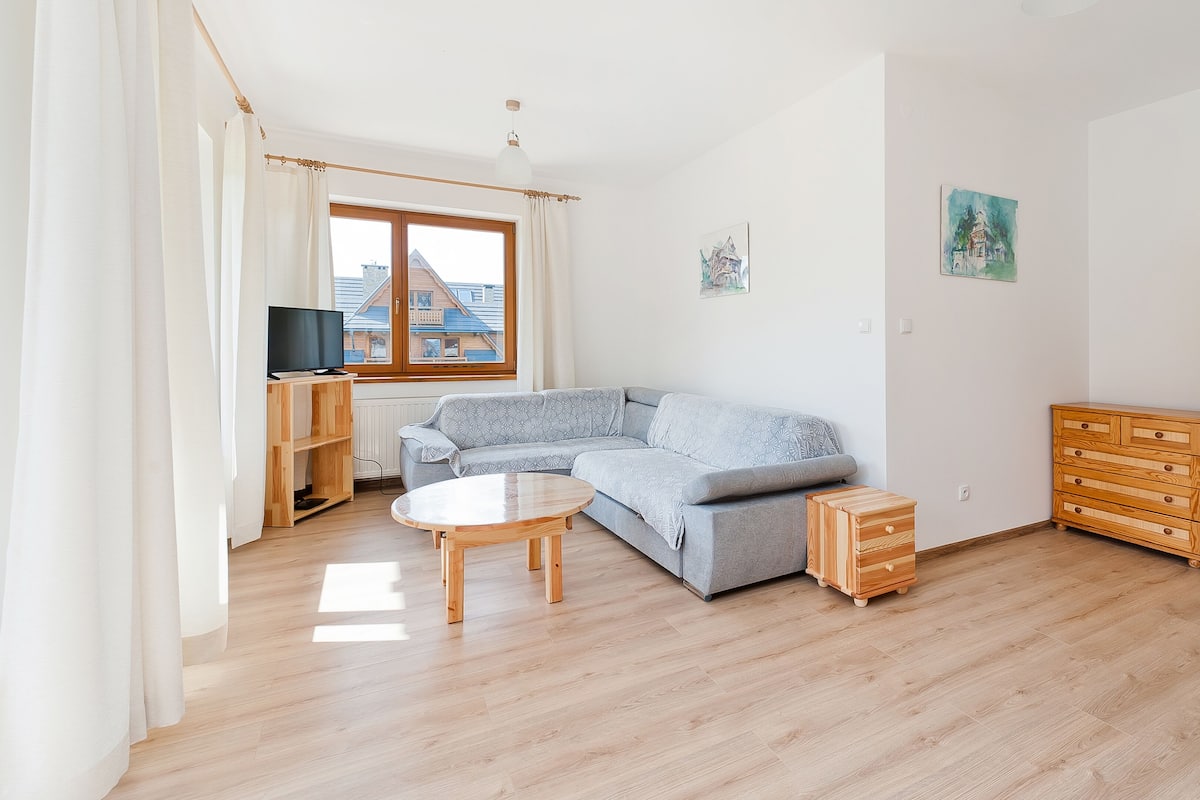 Un salón con un sofá seccional gris, una mesa de centro de madera, un televisor sobre un mueble de madera, un aparador de madera y una ventana con cortinas.