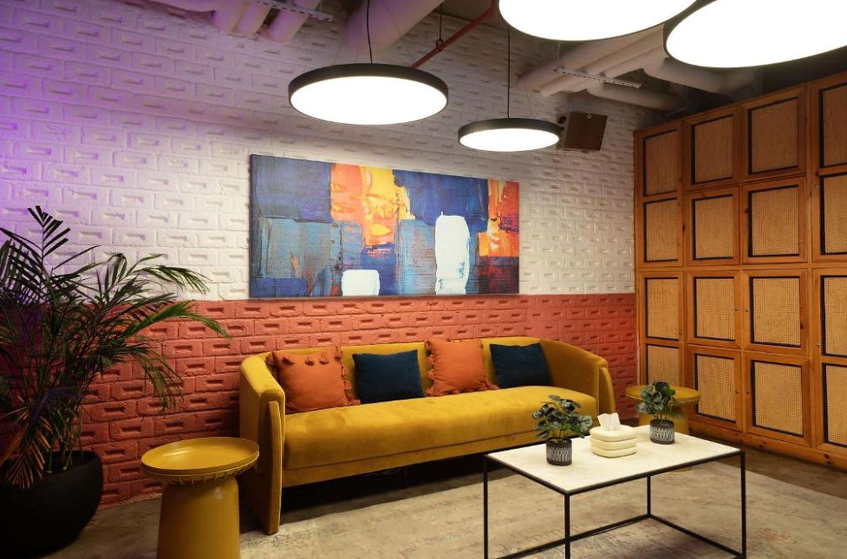 A room with a brick wall, a yellow sofa, a coffee table, and a large abstract painting.