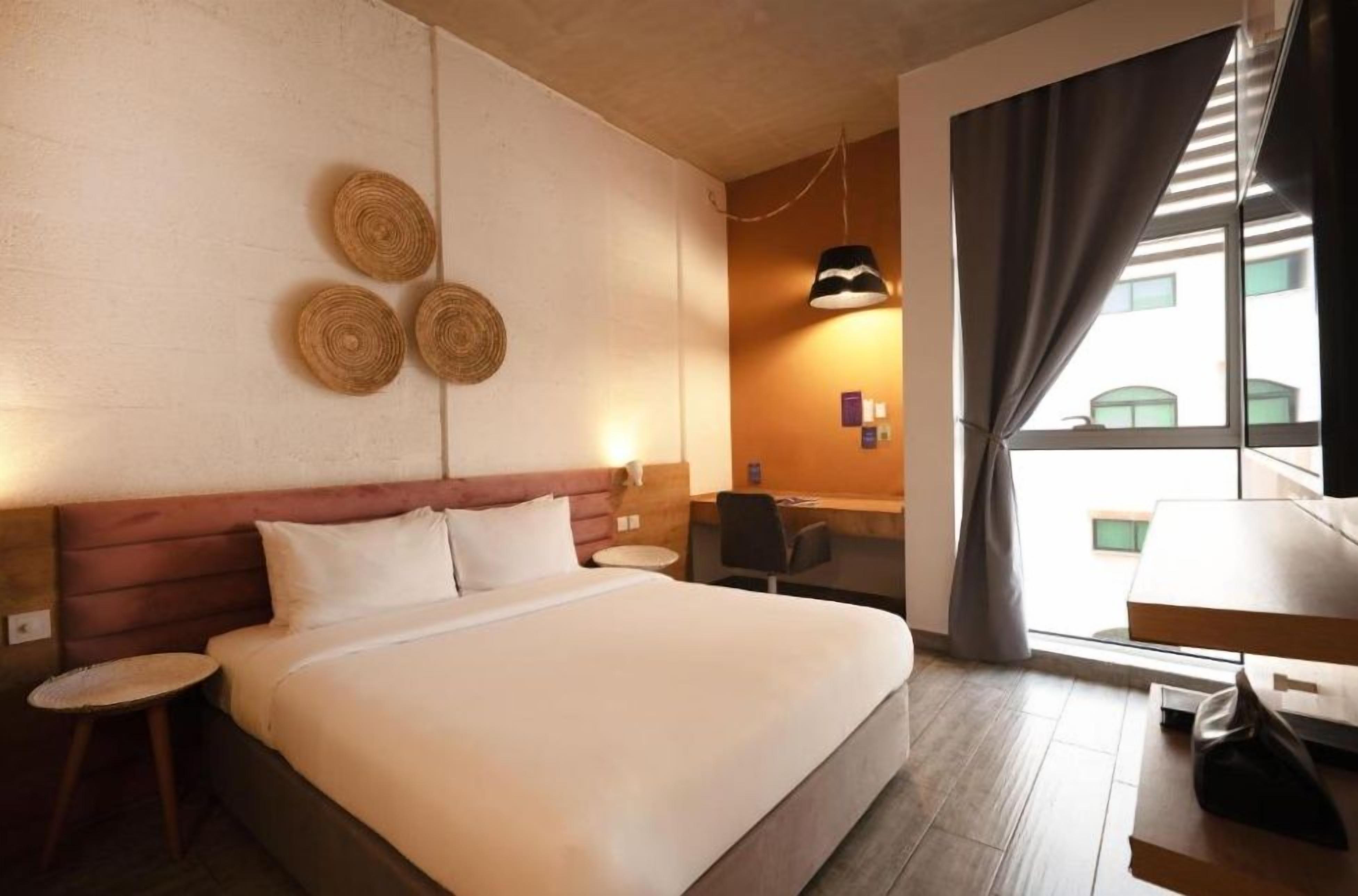 Una habitación de hotel con cama, escritorio, silla, lámpara y ventana con cortinas.