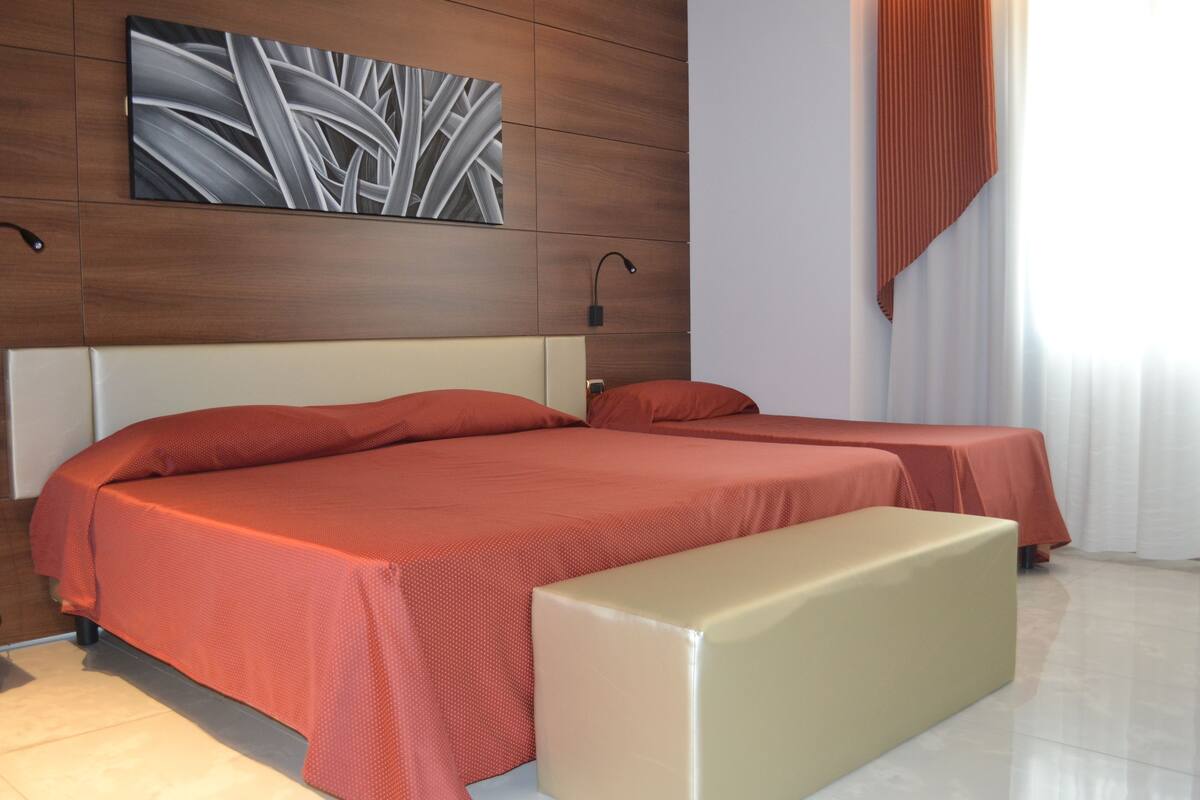 A modern bedroom with two beds, a bench, a wall-mounted artwork, and a nightstand.