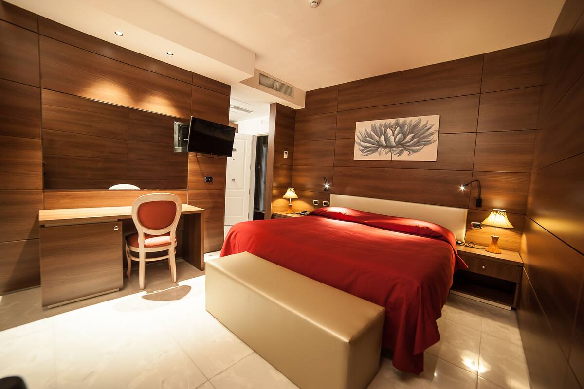 A modern bedroom with a wooden wall panel, a bed with red bedding, a desk with a chair, and bedside lamps.