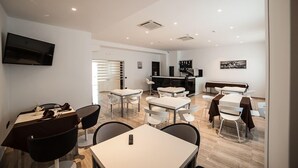A modern dining area with white tables and chairs, a flat-screen TV mounted on the wall, and a kitchen area with appliances. - Family Hotel (Polla)