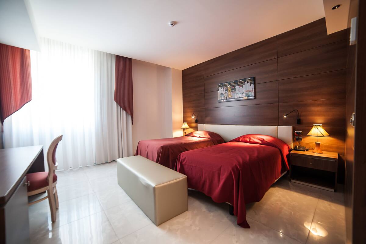 A hotel room with two beds, a desk, a chair, and a bench.