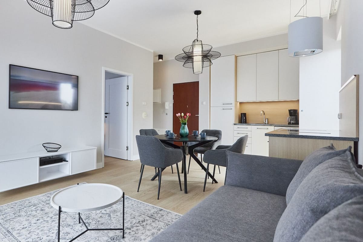 A modern living room with a grey sofa, a dining table with chairs, a TV mounted on the wall, and a kitchen area with white cabinets and a coffee machine.