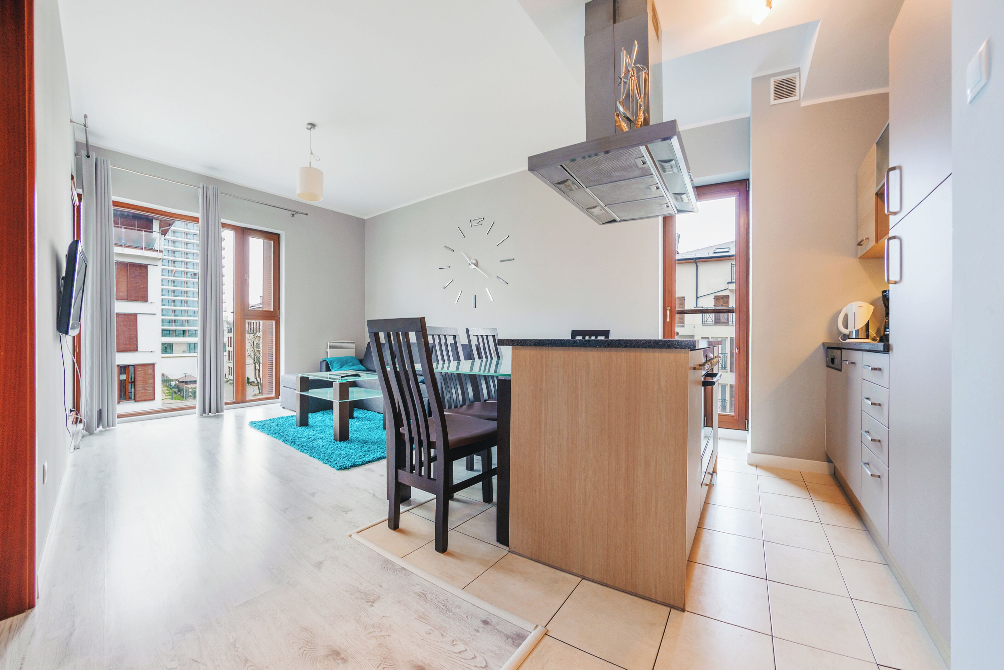 A modern kitchen with a central island, a dining area with chairs, and a large wall clock.