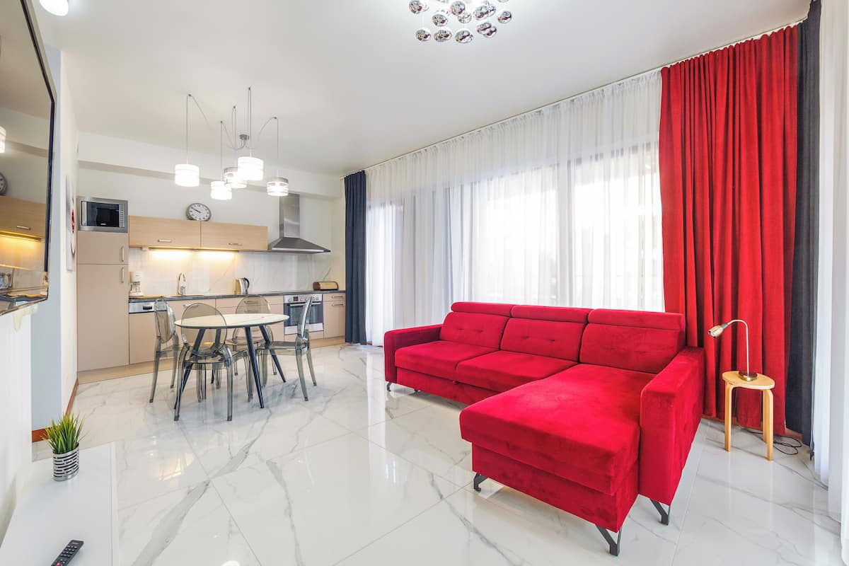 A modern living room with a red sectional sofa, a dining area with a glass table, and a kitchen with white cabinets and stainless steel appliances.