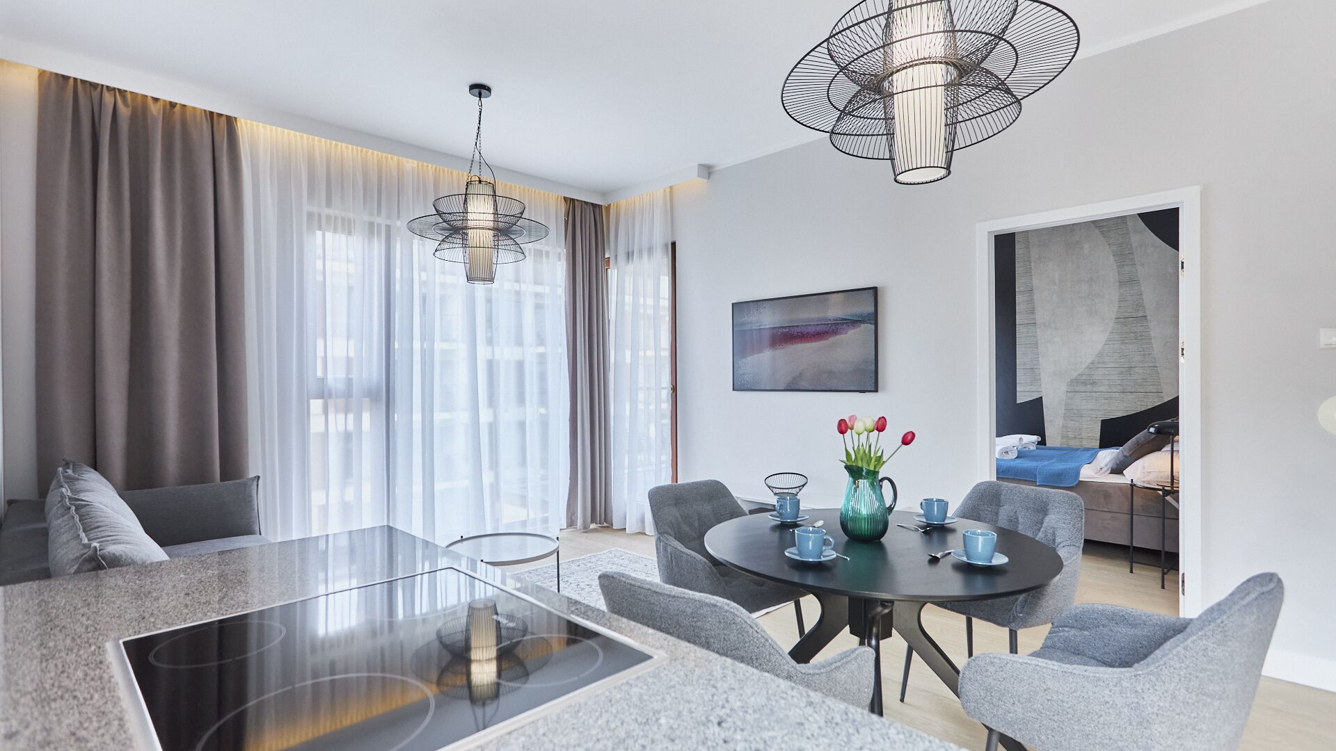A modern living room with a glass dining table, grey sofas, and a large window with curtains.