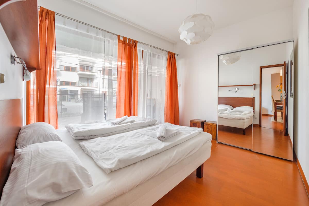 A hotel room with a large bed, a bedside table, a mirror, and a view of the street through sheer curtains.