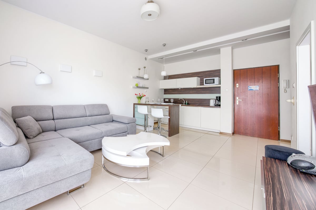A modern living room with a grey sectional sofa, a white coffee table, and a kitchen area with a microwave and a sink.