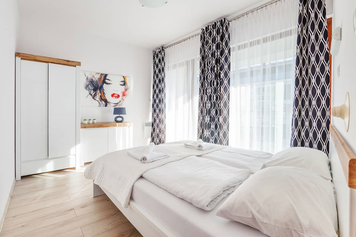 A modern hotel room with a wooden floor, a bed with white linens, a bedside table with a lamp, and a painting on the wall.