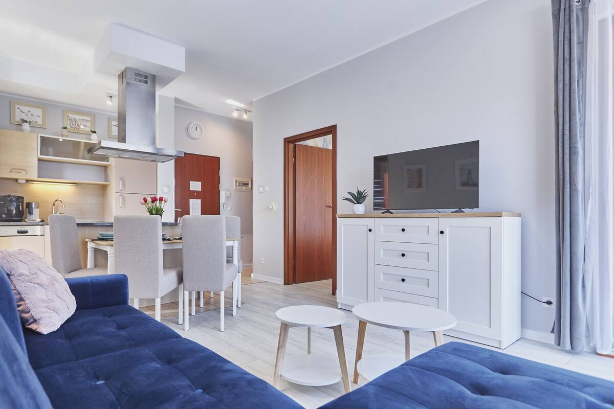 A modern living room with a blue sofa, a dining area with chairs, a kitchen with a stove, and a TV on a cabinet.