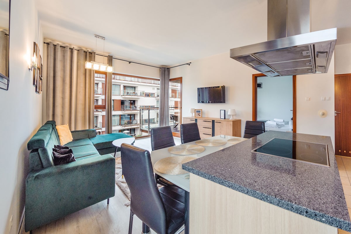 A modern kitchen with a central island, dining area with chairs, a sofa, and a view of the building's exterior.