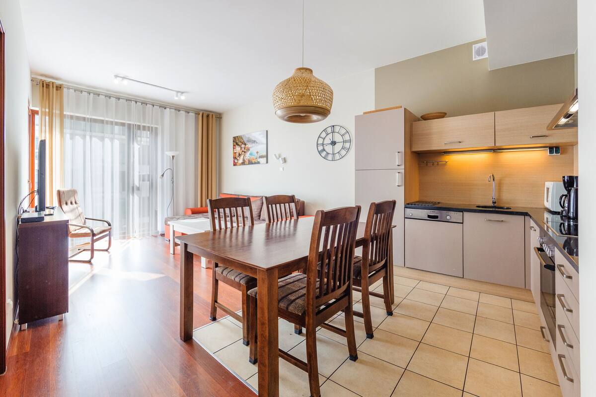 A modern kitchen with a dining area, wooden furniture, and a large window with curtains.