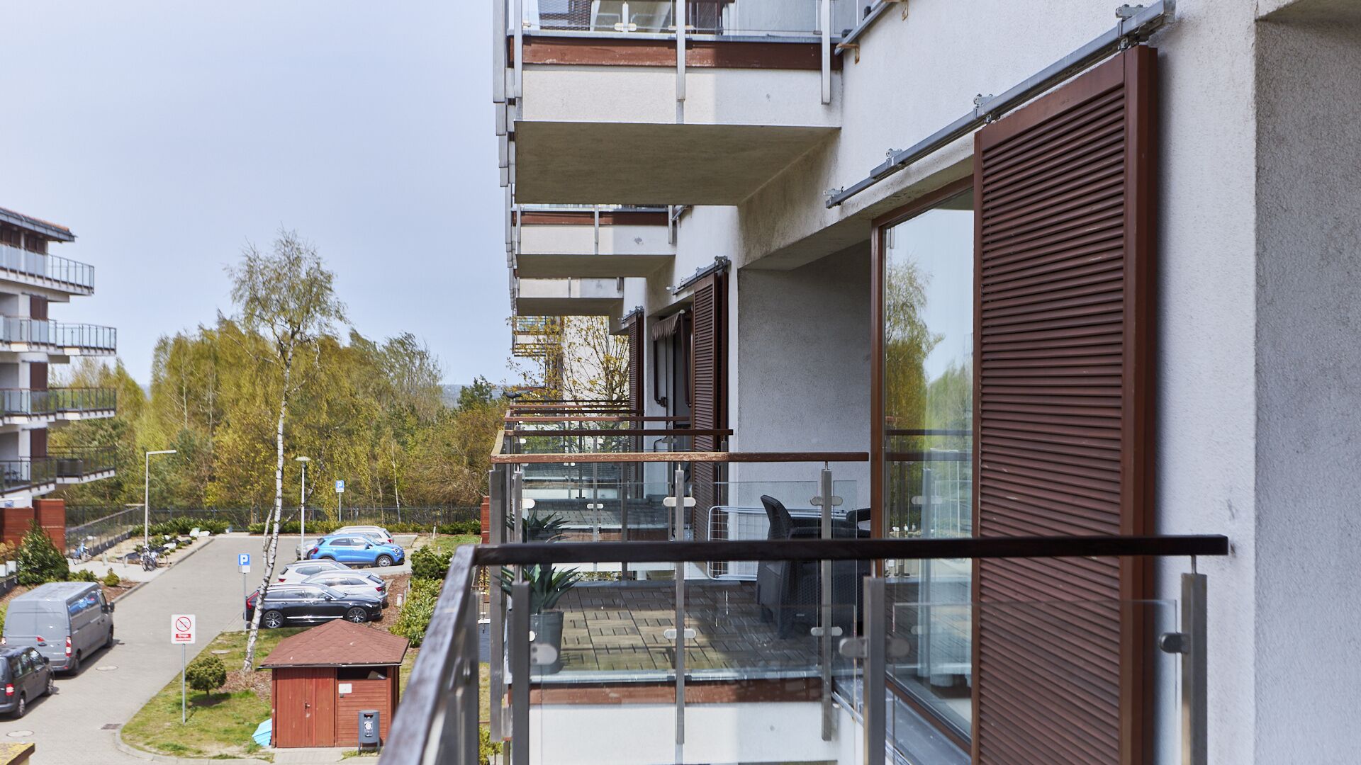 A modern apartment building with balconies, a parking area, and a view of trees and other buildings.