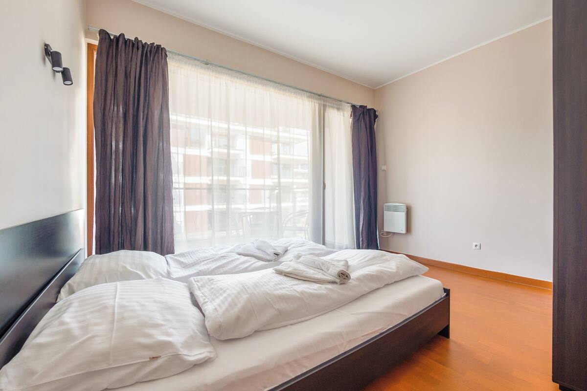 A hotel room with a bed, curtains, a wall-mounted air conditioner, and a window with a view of buildings.