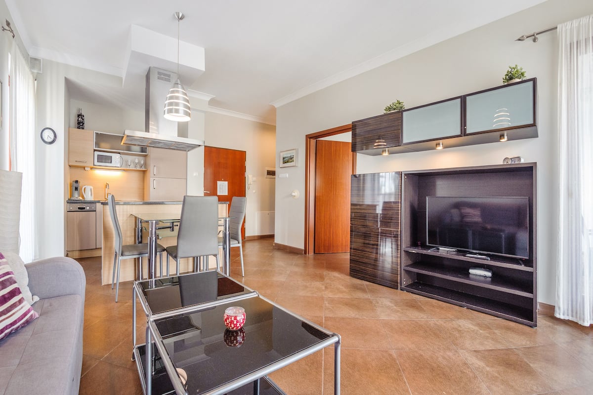 A modern living room with a glass coffee table, a sofa, a dining area with chairs, a kitchen with a microwave, and a TV in a wooden entertainment unit.