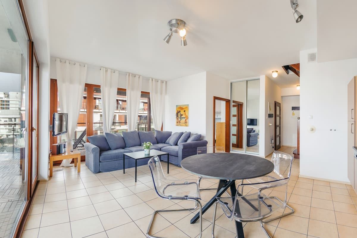 A modern living room with a sectional sofa, a glass dining table, and a kitchen area with a microwave and sink.