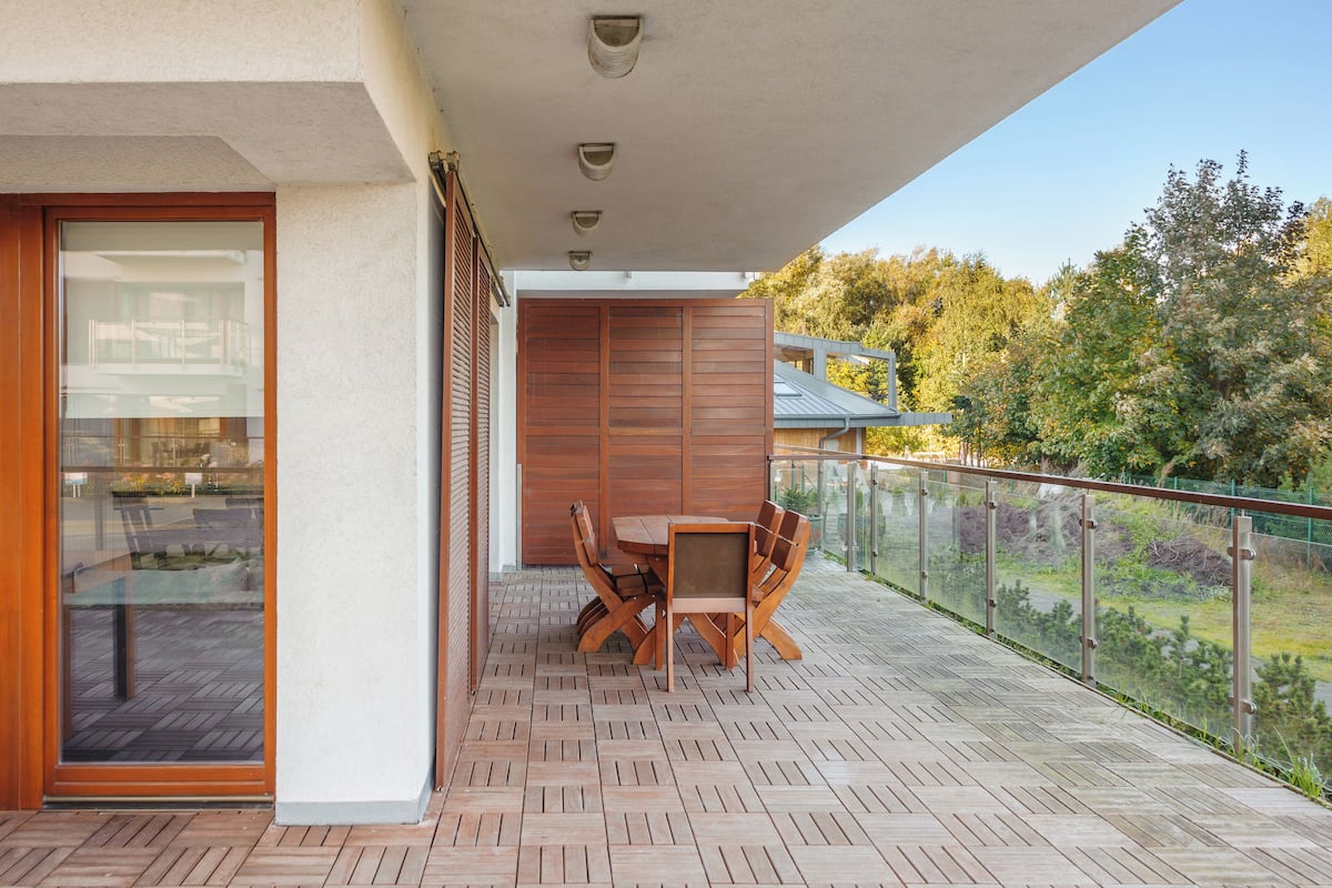 A balcony with a wooden table and chairs, a glass door leading to an indoor area, and a view of trees and a building in the background.