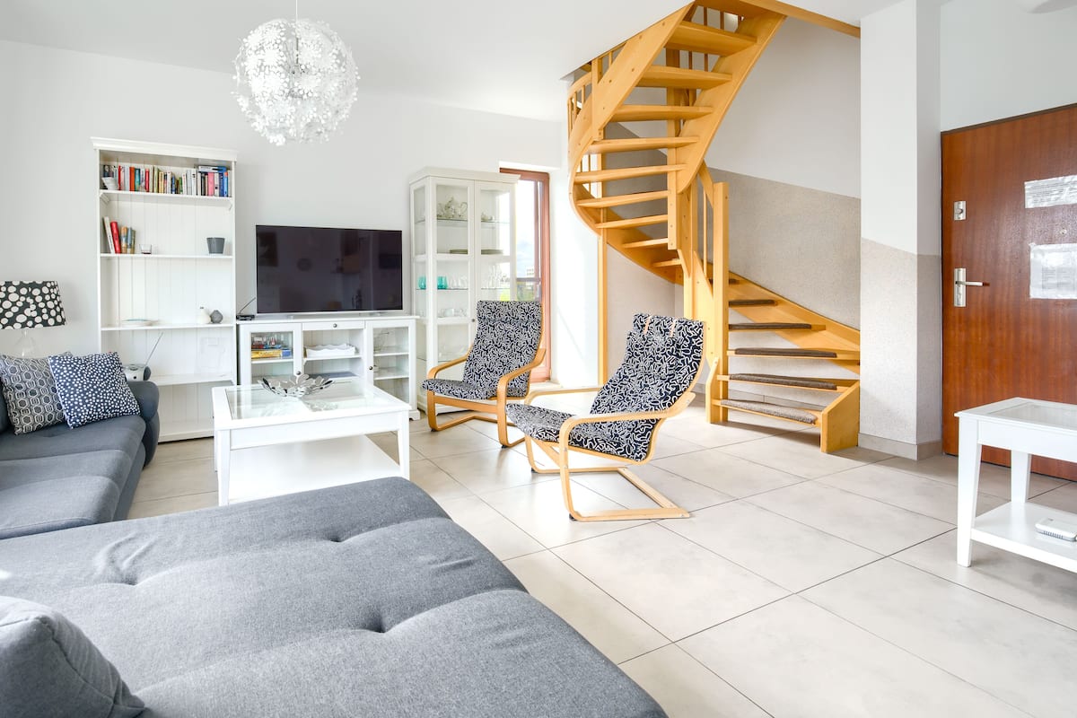 A modern living room with a grey sofa, patterned armchairs, a white coffee table, a TV, and a wooden spiral staircase.