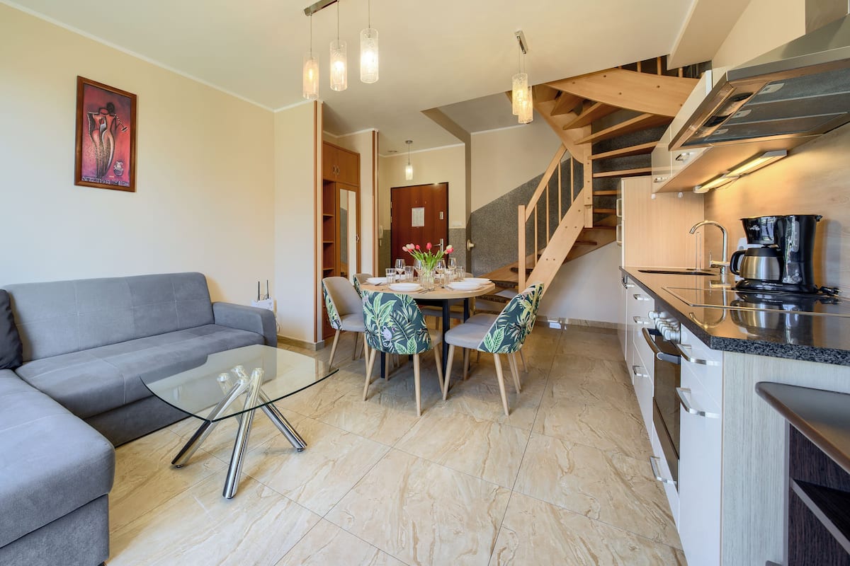 A modern kitchen with a built-in stove, a dining area with a table and chairs, a grey sofa, and a staircase leading to an upper level.
