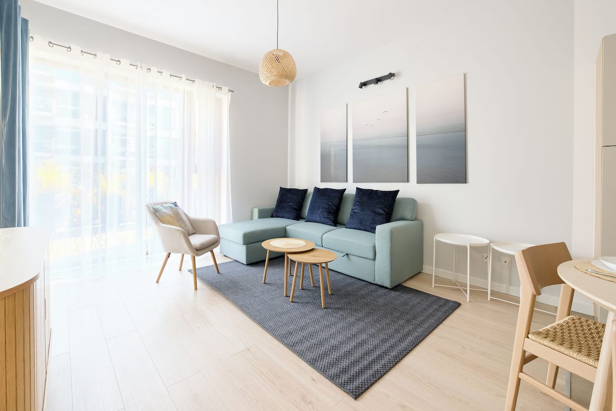A modern living room with a light blue sofa, a round coffee table, a dining table with chairs, and a large window with sheer curtains.