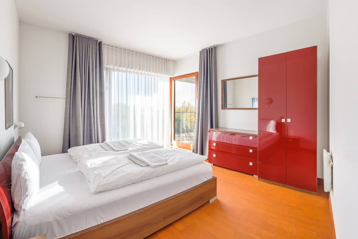 A modern bedroom with a large bed, a red wardrobe, a mirror, and a window with curtains.