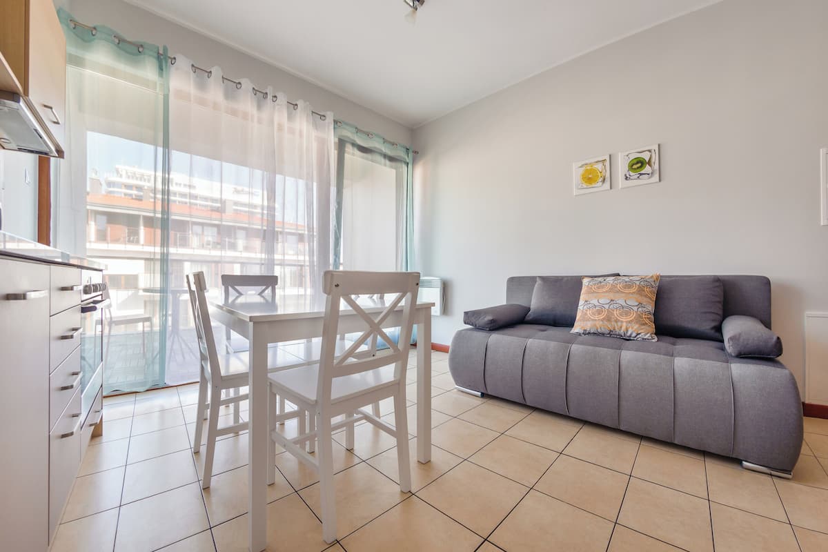 A modern living room with a grey sofa, a white dining table with chairs, and a balcony with a view of buildings.